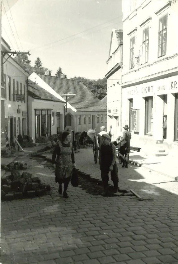 Schwarzweißaufnahme: Leitungsbau in der gepflasterten Schulgasse, Passanten und ein Fuhrwerk